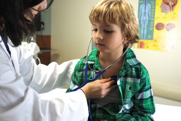 Foto colorida de criança passando por exame de coração, com médica encostando um estetoscópio em seu coração - Metrópoles.