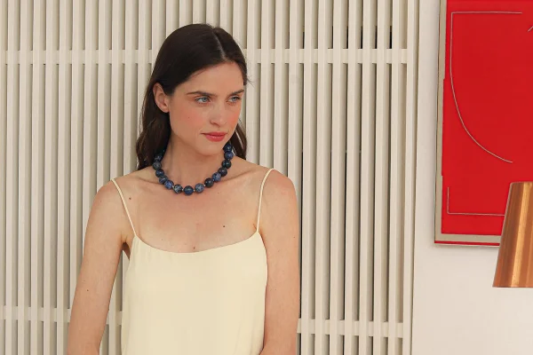 Com painel branco ripado atrás, mulher olha para o lado. Ela usa uma roupa clara com alcinhas e colar azul com pedras esféricas - Metrópoles