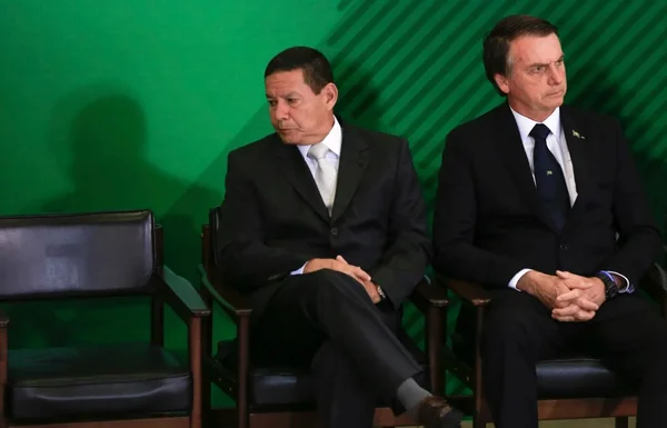 Na imagem colorida, dois homens estão posicionados na direita. Eles usam terno e gravata na cor preta e camiseta branca. Eles tem cabelos curtos e escuros