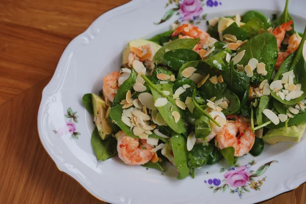 Deliciosa salada de camarões, espinafre e amêndoas servida em um prato decorativo com flores para um aperitivo gourmet. Metrópoles