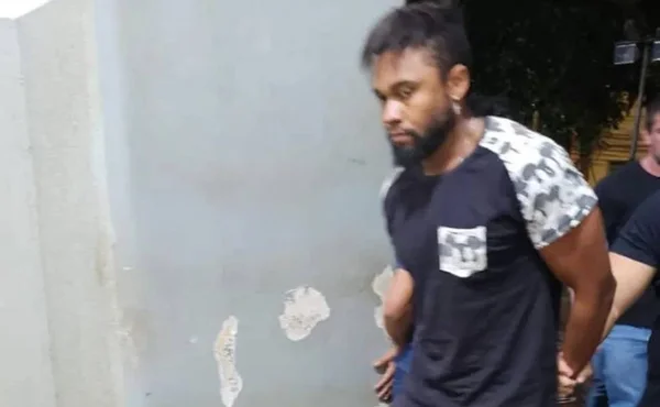 Imagem colorida mostra o cantor sertanejo João Vitor Malaquias sendo conduzido por policiais ao ser preso na cidade de Ribeirão Preto, no interior de São Paulo; ele é um homem negro, com cabelo preto liso e barba, e veste uma camiseta azul escura com bolso estampado - Metrópoles