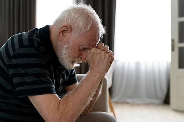 Homem idoso branco sentado no sofá. Ele está inclinado apoiando a cabeça nas mãos em um sintoma de esquecimento, comum em Alzheimer e demência