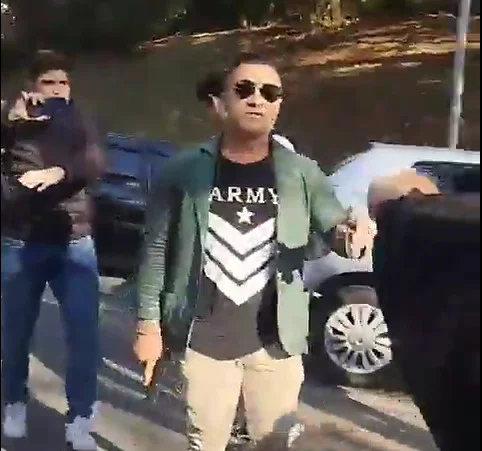 Imagem colorida de homem com camiseta preta, blusa azul e óculos escuros segura revólver apontado para baixo, euquanto sinaliza com outro braço para pessoas se afastarem - Metrópoles