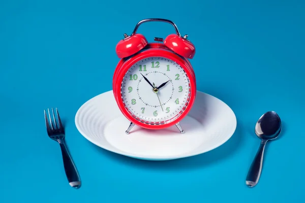 Prato de comida branco com relógio vermelho em cima, talheres ao lado e fundo azul - Metrópoles