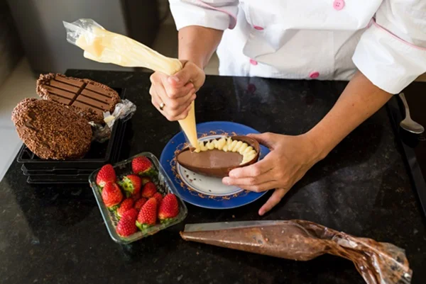 uma confeiteira recheia um ovo de chocolate com creme de baunilha. ela está produzindo o doce em cima de uma mesa com morangos e mais chocolate