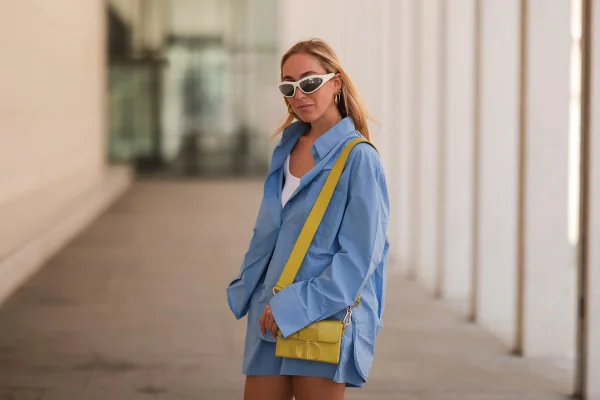 Mulher branca usando uma camisa social com bolsa amarela a tiracolo e óculos branco, de lado - Metrópoles