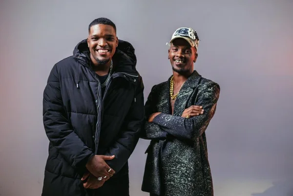 Foto colorida de dois homens negros. Um de casaco preto e outro de boné branco e blusa verde escura - Metrópoles