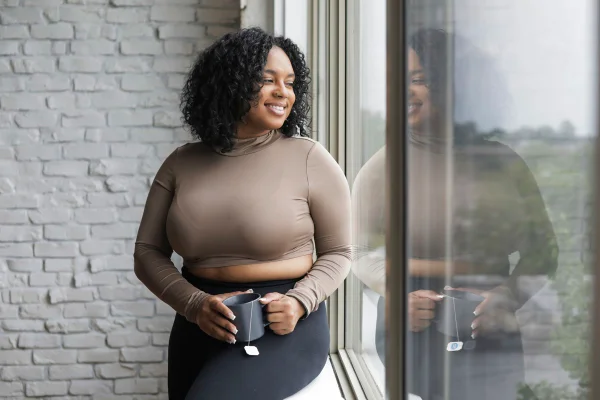 Foto colorida de mulher sentada próximo à janela. Ela segura uma xícara - Metrópoles