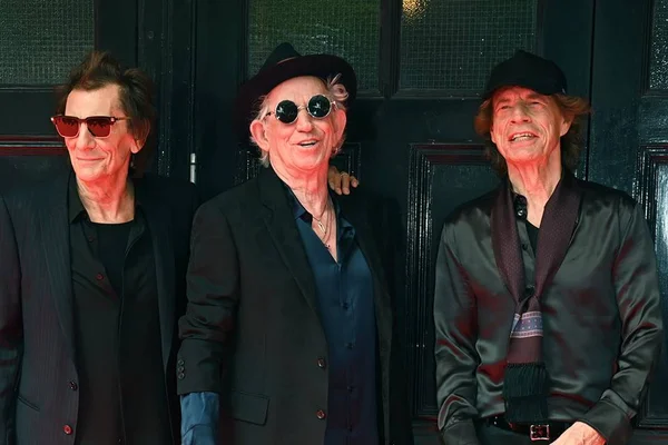 Foto colorida dos integrantes de Ronnie Wood, Keith Richards e Mick Jagger com roupas pretas em fundo preto - Metrópoles