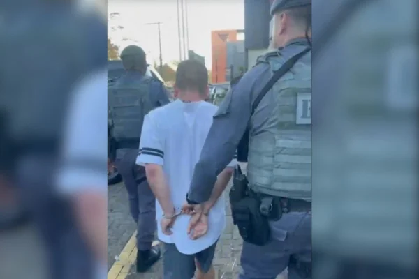 Foto colorida do momento em que suspeito de ligação com o PCC é conduzido por policiais do 1° Batalhão de Ações Especiais (Baep) durante operação do Ministério Público de Santa Catarina.