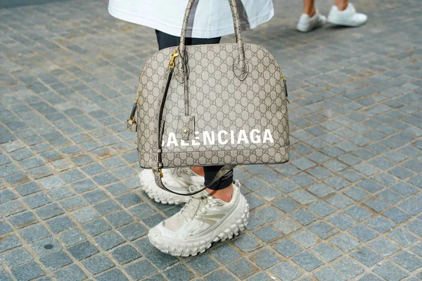 Pessoa andando pelas ruas de Paris com tênis branco, de proporção grande, da marca Balenciaga. Usa ainda uma calça legging preta, uma camiseta branca longa e uma bolsa de couro cinza também da Balenciaga.