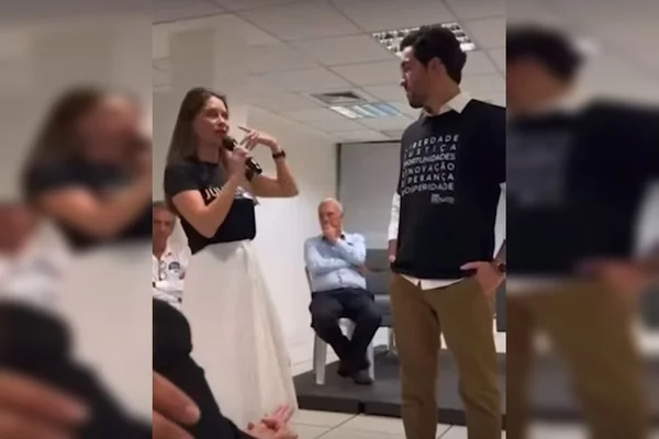 Mulher com camisa preta e saia falando ao microfone e olhando para homem ao lado dela. Ele está em pé, com camisa preta e calça marrom e com as mãos no bolso. Outros homens aparecem sentados ao fundo da sala