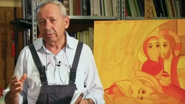 Padre Marko Rupnik de camisa branca e macacão escuro com uma obra sacra ao fundo