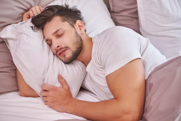 Foto colorida de homem vestindo uma blusa branca, deitado de lado em uma cama, enquanto abraça o travesseiro - Metrópoles.