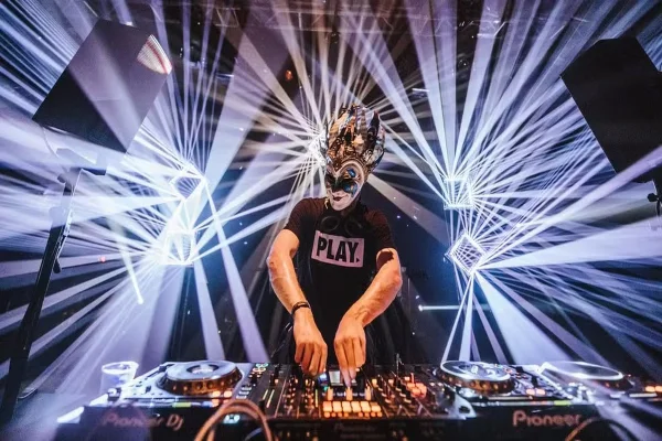 DJ mascarado toca em mesa de DJ com luzes ao fundo