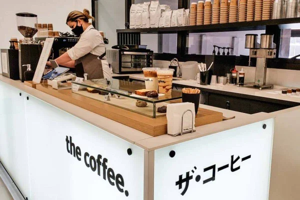 A foto mostra a bancada do The Coffee com atendente fazendo café em uma máquina de espresso