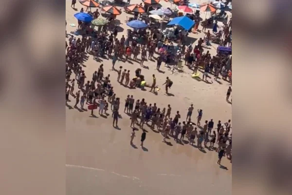 imagem colorida mostra população observando pessoas fazerem manobra de reanimação em banhista na faixa de areia de uma praia