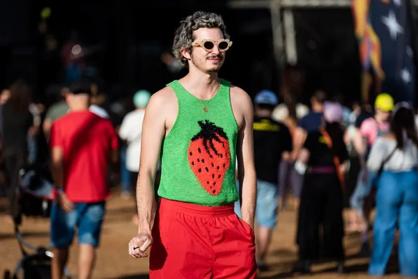 Homem branco e jovem, de cabelo liso descolorido, posando para foto no Festival CoMA, em Brasília. Ele usa um top de tricô verde com um morango grande estampado no centro, uma calça vermelha e um óculos escuro com lentes em formato de flor
