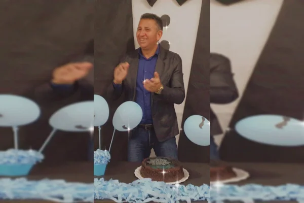 Homem de cabelos grisalhos veste camisa azul escura, blazer e calça jeans. Ele celebra o aniversário. Na frente um bolo, docinhos e balões
