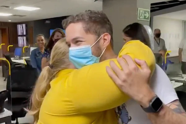 Jornalista Gabriel Luiz volta à Globo em Brasília após ser esfaqueado em assalto e é aplaudido por colegas na redação. Na foto, ele aparece de máscara abraçando mulher loira de camisa amarela - Metrópoles