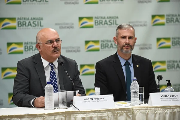 O ex-ministro da Educação ao lado do recém nomeado a cargo, Victor Godoy, na época secretário. Eles falam em coletiva de imprensa sentado frente a mesa com microfones - Metrópoles