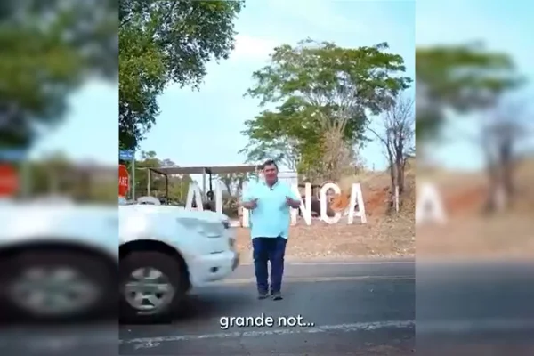 Imagem colorida de um homem de camisa azul claro e calça azul escuro sendo quase tocado por um carro