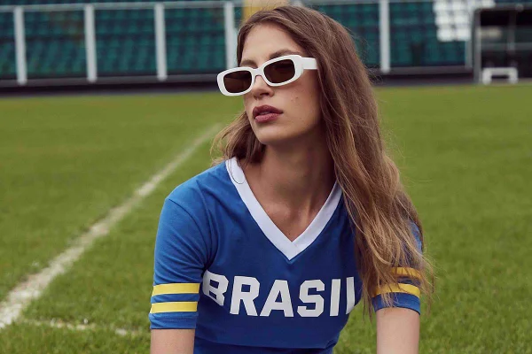 Mulher branca e jovem, com cabelos longos, lisos e loiros, posando para foto sentada no chão do gramado de um campo de futebol. Usa um óculos escuros com armação branca e uma camiseta azul com a palavra BRASIL escrita em branco e listras amarelas na manga.