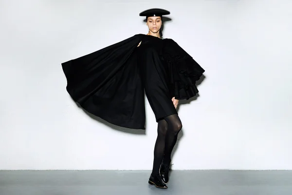 Mulher branca posando para foto em parede branca. Ela usa um vestido preto com mangas gigantes, além de um chapéu e meia calça, tudo também na cor preta.