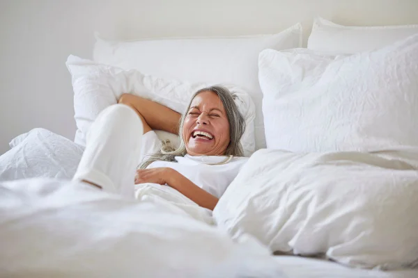 Foto mostra mulher de meia idade e cabelos grisalos deitada em uma cama de lençóis brancos. Imagem ilustrativa sobre prazer após a menopausa