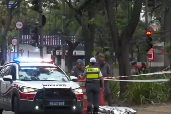 Carros da Polícia Militar ao lado de canteiro central de avenida isolado por fitas; um corpo está coberto por manta de alumíniio - Metrópoles