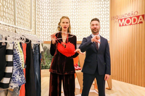 Os apresentadores Renata Kuerten e Lucas Anderi, do programa Esquadrão da Moda, posa para foto em cenário. Ela é uma mulher branca e jovem, de cabelo liso loiro, e usa um terno marrom de veludo. Ele é um homem branco e jovem, de cabelo e barba grislha, e usa terno completo cinza. - Metrópoles