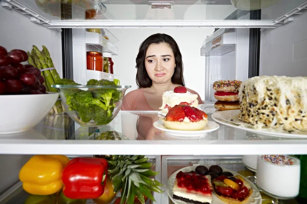 Foto colorida de mulher branca com a cabeça dentro da geladeira, cheia de bolos, doces, legumes e frutas - Metrópoles