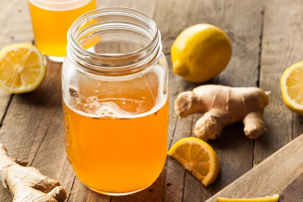 Foto colorida de kombucha. Imagem mostra um recipiente com um líquido alaranjado e vários limões e gengibre do lado - Metrópoles