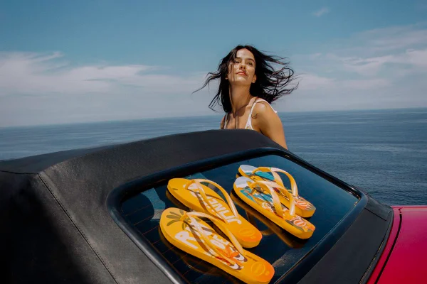 Em praia, com mar ao fundo, mulher com cabelos ao vento. À frente um carro com sandálias amarelas da Havaianas no capô - Metrópoles