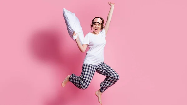 Mulher de pijama com travesseiro na mão pulando em frente a fundo rosa