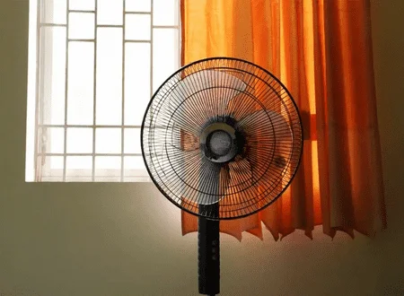 Fotografia colorida mostra ventilador na frente de janela de quarto com uma cortina laranja no lado esquerdo