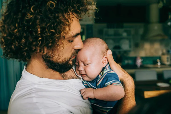 Homem branco com cabelo enrolado segurando bebê e cheirando sua cabeça