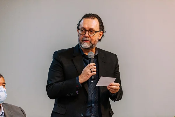 Fotografia colorida mostra Carlos Bezerra Júnior, da cintura para cima, um homem de barbas grisalhas, óculos redondo, vestindo terno preto e segurando um microfone com a mão direita e um papel com a esquerda - Metrópoles