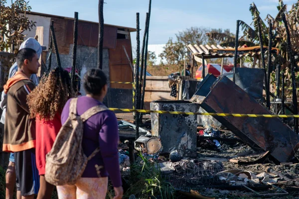 Imagem colorida de pessoas vestidas com roupas de cores diversas, em frente a um barraco deteriorado por um incêndio. Há uma faixa preta e amarela em frente