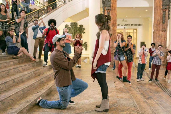 foto colorida de homem ajoelhado segurando anel e mulher em pé sorrindo e pessoas ao fundo olhando - metrópoles