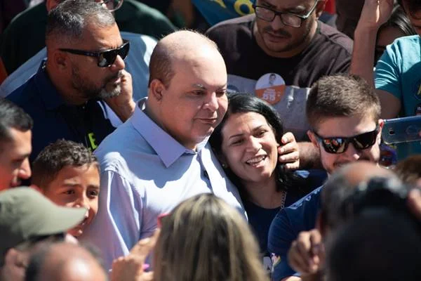 Fotografia colorida de homem de camisa abraçando mulher com pessoas ao redor