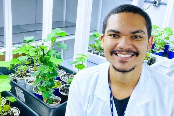 Imagem colorida de um homem preto de barba e bigode, usando um jaleco em frente a alguns vasos de plantas