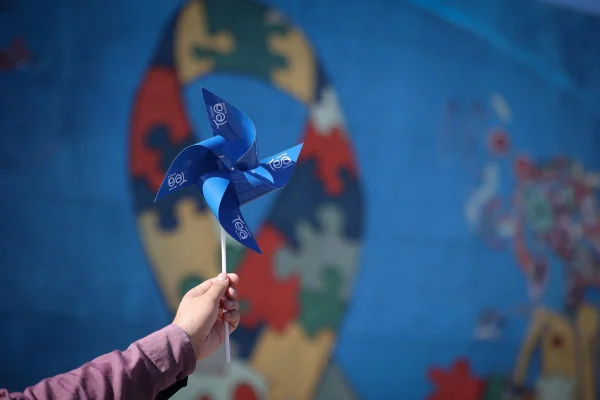 Imagens coloridas mostram a mão de uma criança segurando um catavento azul com a inscrição TEA e o símbolo do autismo ao fundo, pintado em um muro azul