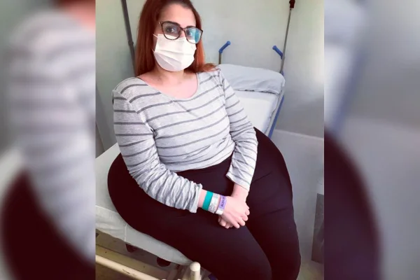 Imagem colorida com a mulher Karina Rodine sentada em uma cama. Karine vive em Curitiba, no Paraná, e tem um tumor de 40Kg