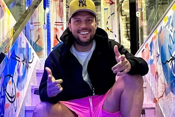Thiago Servo posa de moletom preto e boné amarelo, sentado em uma escada - Metrópoles
