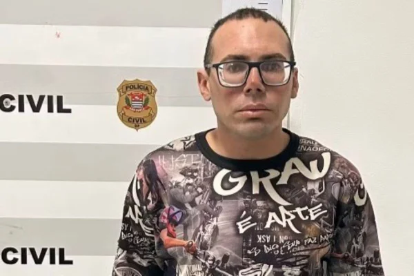 Homem careca de óculos com camiseta estampada no momento da prisão. Ao fundo, logo da policia civil de sao paulo; ele foi preso acusado de feminicídio - Metrópoles