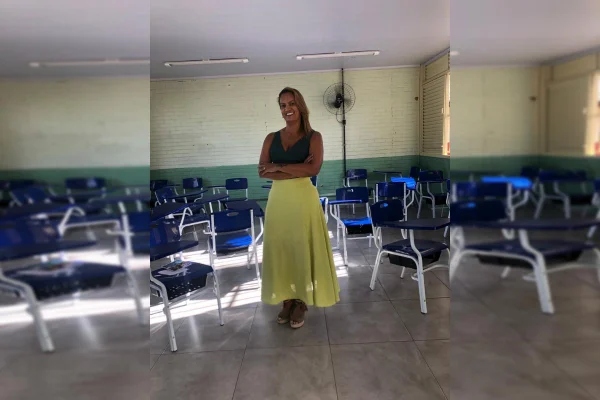 Mulher de braços cruzados, dentro de sala de aula, com blusa verde e saia amarela
