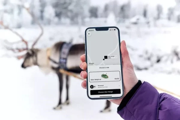Foto colorida de uma rena em um fundo de neve e na frente um celular aberto no aplicativo Uber