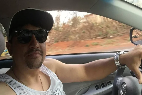 Foto colorida de homem com boné e óculos dirigindo um carro