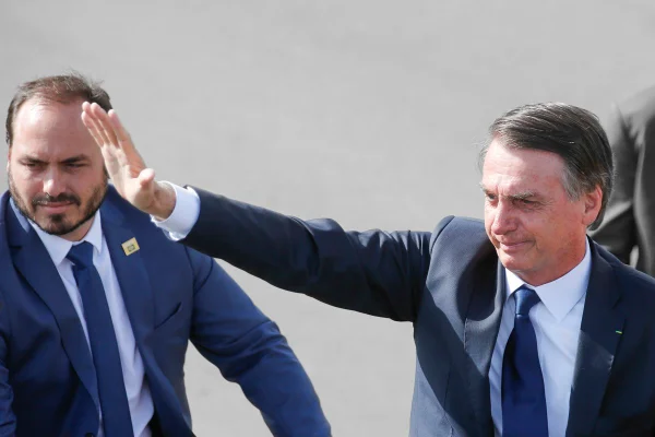 O ex-presidente Bolsonaro acena em evento ao lado de seu filho, Carlos Bolsonaro. Ambos sorriem e usam terno - Metrópoles
